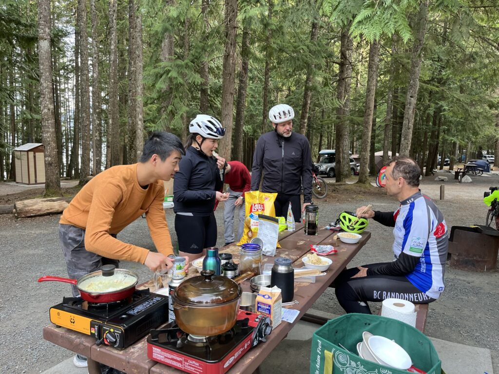 Mirtha Gamiz, Jonathan Hanvelt, and Craig Premack at a picnic table. Fabian Poon is preparing food.
